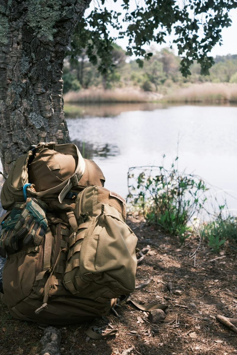 tactical backpack sitting on ground near a tree and lake